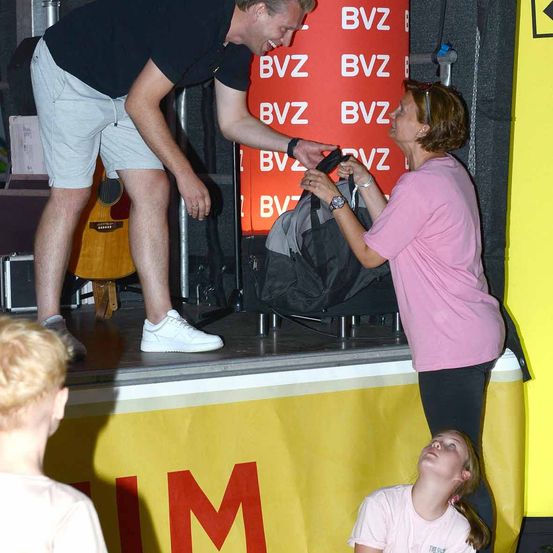 Ein Mann auf der Bühne gibt einer Frau eine Tasche, während ein Mädchen zu ihnen aufblickt. Ein gelber Banner mit roten Buchstaben ist im Hintergrund.