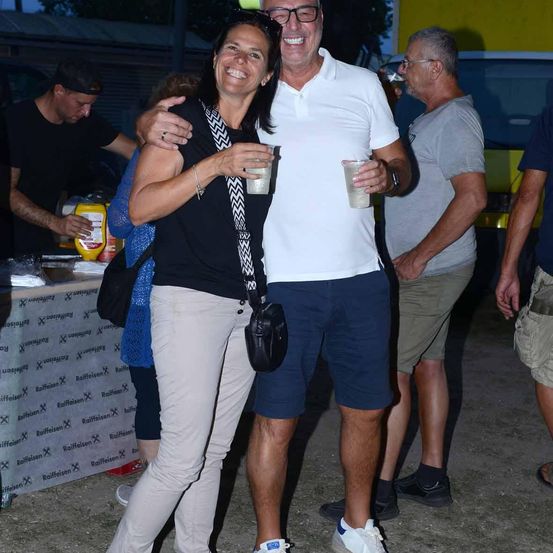 Eine Gruppe von Menschen steht und lächelt, ein Mann und eine Frau halten Getränke, ein weiterer Mann mit Brille hinter ihnen und ein Tisch mit Essen im Hintergrund.