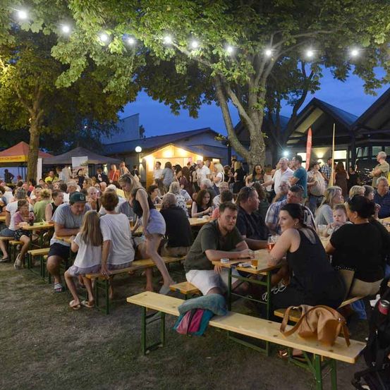 Eine große Gruppe von Menschen versammelt sich für eine Outdoor-Veranstaltung bei Nacht, sitzt um Tische unter Bäumen mit Lichterketten.