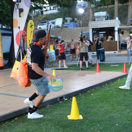 Ein Mann läuft auf einer Bühne und hält einen Ball in der Hand, mit orangefarbenen Kegeln auf dem Rasen. Hinter ihm stehen und gehen Leute umher, und ein Imbisswagen ist an der Seite geparkt.