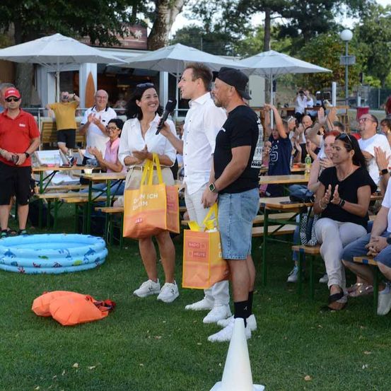 Eine Gruppe von Menschen ist im Freien versammelt, einige stehen und andere sitzen auf Bänken. Zwei Personen halten orange Taschen und stehen vor einem Mikrofon. Die Umgebung umfasst Tische, Schirme und einen kleinen aufblasbaren Pool.