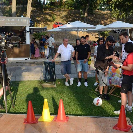 Eine Gruppe von Menschen versammelt sich auf einer Rasenfläche, möglicherweise für ein Fotoshooting. Ein Mann in einem weißen Hemd geht auf einen Mülleimer zu. Es gibt Verkehrskegel und einen Fußball auf dem Boden. Eine Kamera steht auf einem Stativ, und ein Imbisswagen ist im Hintergrund.