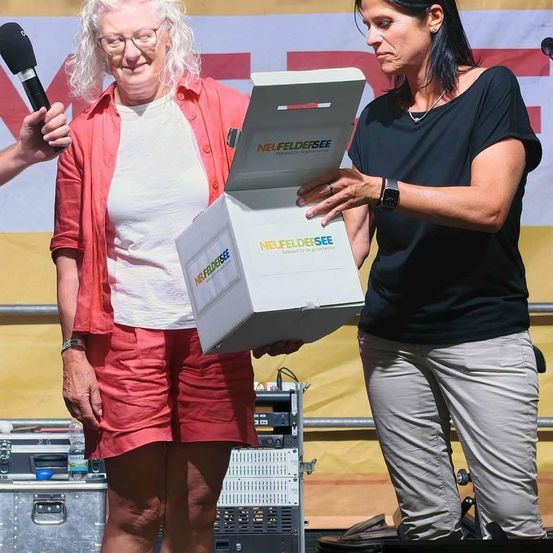 Zwei Frauen auf der Bühne, eine hält eine Box mit der Aufschrift 'NEUFELDSEE', die andere lächelt und schaut auf die Box. Ein Mikrofon wird von einer anderen Person hinter ihnen gehalten.
