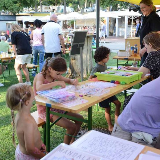 Kinder und Erwachsene sitzen draußen an Tischen und malen Mandalas aus. Eine Frau in Schwarz steht hinter ihnen und lächelt. Im Hintergrund gehen und sitzen Leute unter Schirmen an Tischen.