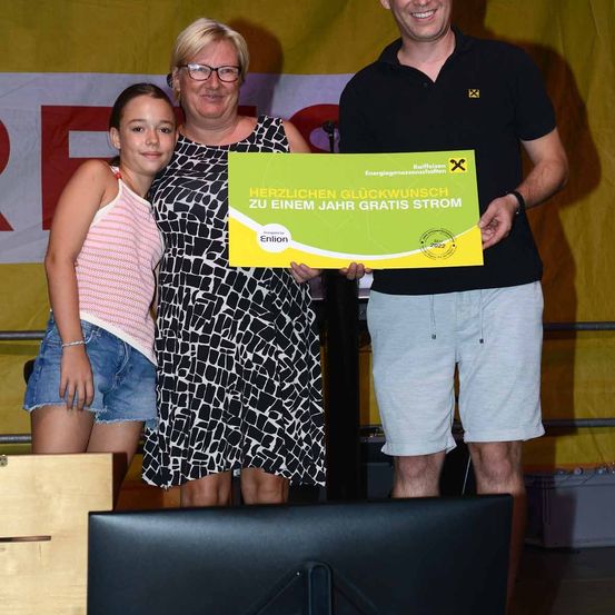 Drei Personen stehen auf einer Bühne, lächeln, wobei eine Frau ein grünes Zertifikat hält, das 'Herzlichen Glückwunsch zu einem Jahr gratis Strom' besagt. Die Frau trägt eine Brille und der Mann ein schwarzes Hemd. Ein Mädchen steht links.