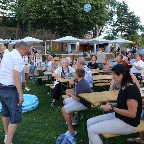 Eine Gruppe von Menschen versammelt sich in einer Freiluftumgebung, möglicherweise in einem Park. Einige sitzen an Tischen, während andere stehen und interagieren. Ein Mann in einem weißen Hemd schaut auf eine schwebende Blase. Das Gebiet ist von Bäumen und Sonnenschirmen umgeben.