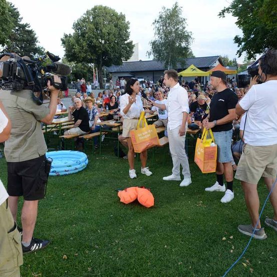 Eine Gruppe von Menschen versammelt sich im Freien, wobei zwei Männer orangefarbene Taschen halten. Eine Frau in der Mitte spricht zu ihnen. Dahinter sitzt eine Menschenmenge an Tischen, und zwei Personen filmen mit Kameras. Bäume und ein Gebäude sind im Hintergrund.