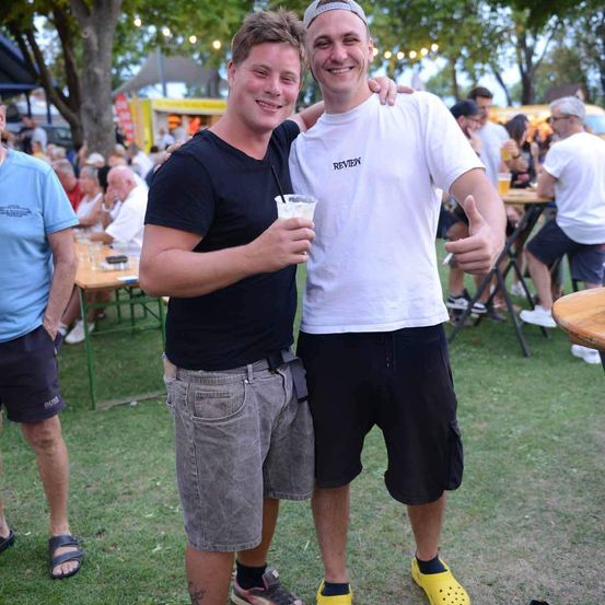 Zwei Männer stehen bei einer Outdoor-Veranstaltung zusammen, einer hält ein Getränk, beide lächeln und geben ein Daumen hoch.