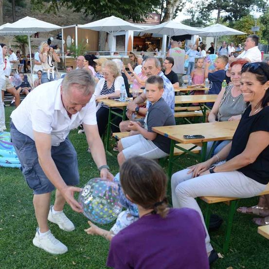 Eine Gruppe von Menschen versammelt sich im Freien, einige sitzen an Picknicktischen. Ein Mann beugt sich vor, um einem Kind einen bunten Luftballon zu geben.