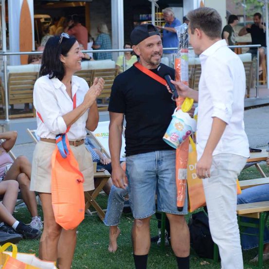 Ein Mann und eine Frau stehen und unterhalten sich, während sie Taschen halten. Hinter ihnen sitzen Leute auf Bänken und Stühlen. Der Mann trägt ein schwarzes T-Shirt, Jeansshorts und eine Kappe.