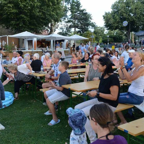 Eine Gruppe von Menschen sitzt an Tischen in einem Außenbereich, möglicherweise in einem Park. Einige klatschen, während andere in Gespräche vertieft sind. Der Bereich ist von Bäumen und Sonnenschirmen umgeben.