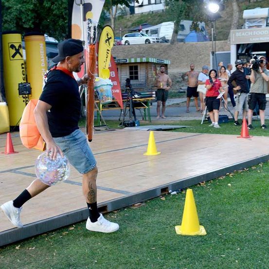 Ein Mann in einem schwarzen T-Shirt und Jeansshorts läuft auf einer Bühne mit einer großen Seifenblasenkugel, umgeben von gelben Kegeln. Im Hintergrund stehen Menschen und schauen zu, ein weißes Auto ist auf einem Hügel geparkt.