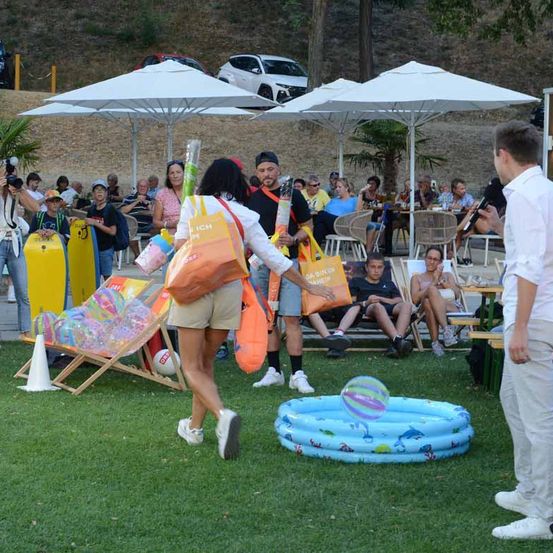 Eine Frau mit Taschen geht an einem Mann mit Mikrofon vorbei, in der Nähe eines kleinen Pools und Strandstühlen, mit Menschen unter Sonnenschirmen im Hintergrund.