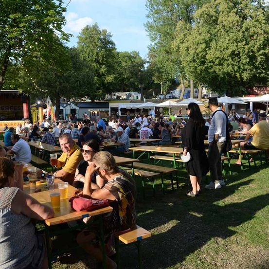 Eine große Gruppe von Menschen sitzt an Picknicktischen in einem Freiluftpark und genießt Essen und Getränke. Bäume und Schirme spenden Schatten.