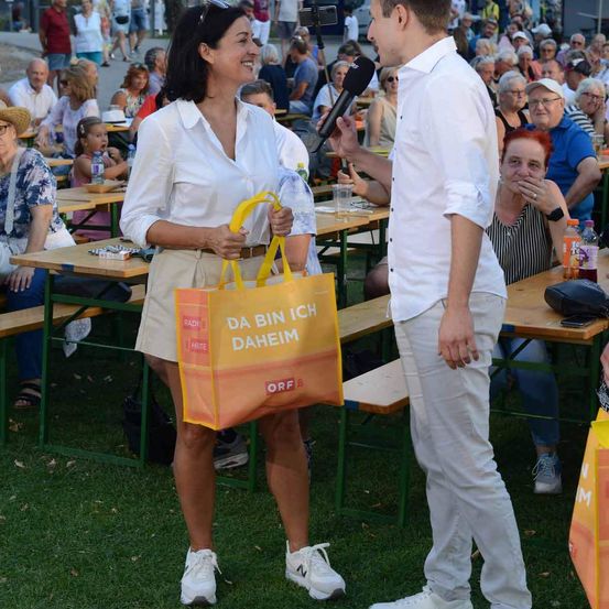 Eine Frau mit einer gelben Tasche, auf der 'Da bin ich daheim' steht, steht vor einem Mikrofon, das von einem Mann gehalten wird, umgeben von einer Menge, die an Tischen sitzt.