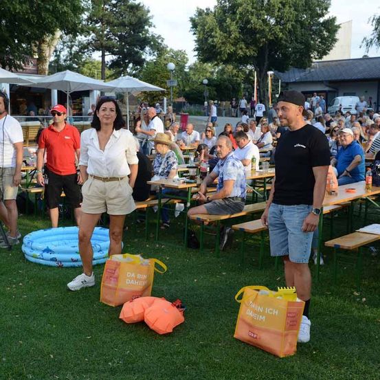 Eine Gruppe von Menschen versammelt sich in einem Außenbereich, wahrscheinlich für eine Veranstaltung. Eine Frau und ein Mann stehen vorne und halten Taschen. Hinter ihnen sitzen Menschen auf Bänken, einige mit Getränken. Bäume und ein Gebäude sind im Hintergrund.