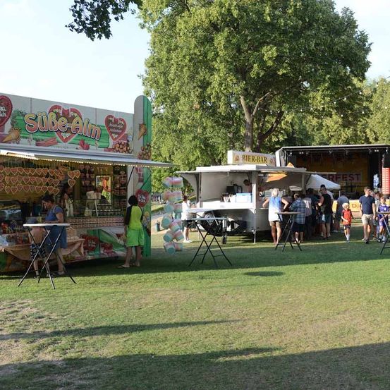 Ein Food-Truck-Festival mit verschiedenen Ständen, darunter einer mit der Aufschrift 'Sue-Alm', ist in einem Park aufgebaut, wo die Leute die Veranstaltung im Freien genießen.