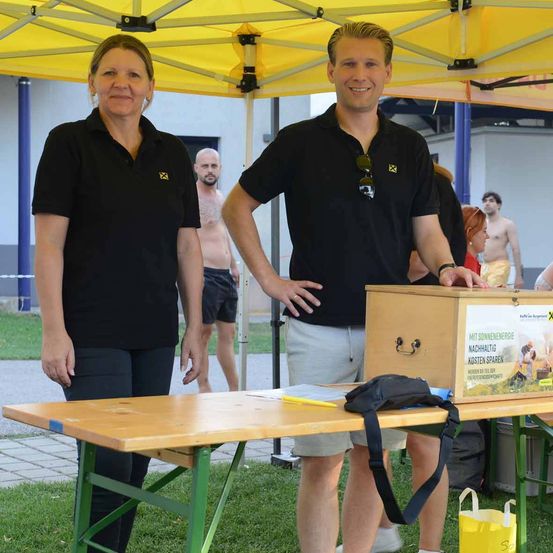 Ein Mann und eine Frau stehen unter einem Zelt, lächeln und posieren für ein Foto. Der Mann hält eine Holzkiste mit einem gelben Etikett. Ein Tisch mit einer Tasche und einem gelben Beutel steht vor ihnen. Im Hintergrund stehen Leute in der Nähe eines Gebäudes.