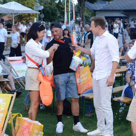 Ein Mann in einem schwarzen T-Shirt und Shorts schüttelt die Hand einer Frau in einem weißen Hemd und beige Shorts. Sie stehen auf einem Rasenfeld, während ein Mann in einem weißen Hemd und Hose ein Mikrofon hält und spricht. Es gibt Menschen, die auf der Seite sitzen und stehen. Es gibt auch Tische und Stühle mit Sonnenschirmen.