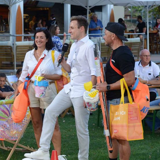 Ein Mann mit einem Mikrofon interviewt eine Frau in einem weißen Hemd und Khaki-Shorts. Sie stehen auf einer Rasenfläche, im Hintergrund sitzen Menschen auf Stühlen und Tischen. Ein weiterer Mann in einem schwarzen T-Shirt hält eine gelbe Tasche und ein Mikrofon.