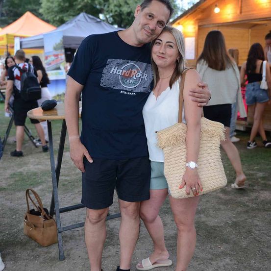 Ein Mann und eine Frau stehen eng beieinander auf einer Outdoor-Veranstaltung und lächeln. Der Mann trägt ein Hard Rock Cafe T-Shirt, und die Frau hält eine Strohtasche. Hinter ihnen gehen Leute an Ständen und Zelten vorbei.