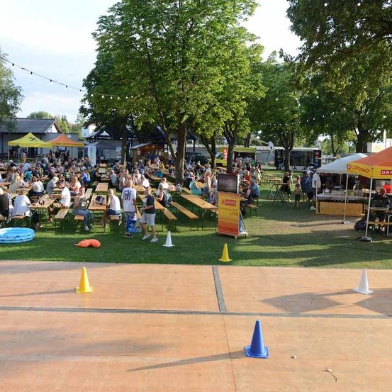 Ein großer Außenbereich mit vielen Menschen, die an Picknicktischen unter Zelten sitzen, umgeben von Bäumen und Grün. Es gibt Food Trucks und eine Bühne mit einem Banner.