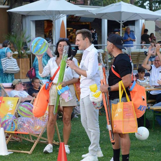 Drei Personen stehen auf einem Rasenfeld mit Strandbällen und Strandspielzeug. Eine Frau hält einen Strandball und ein Mikrofon, während ein Mann einen Strandball und einen Strandschirm hält. Ein anderer Mann hält einen Strandball und eine Tasche.