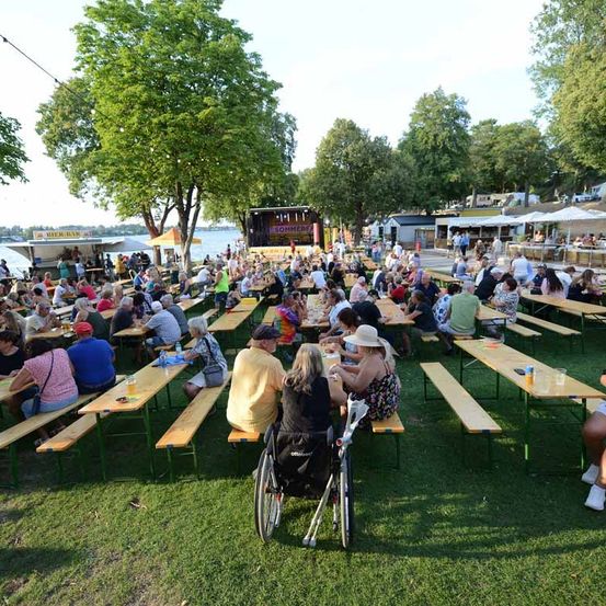 Eine große Gruppe von Menschen sitzt an Tischen auf einer Rasenfläche, wahrscheinlich bei einem Festival. Im Hintergrund befinden sich Bäume, eine Bühne und ein See.