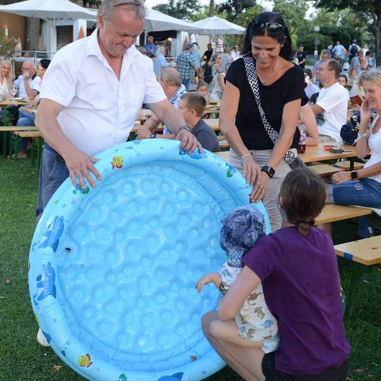 Eine Familie spielt mit einem aufblasbaren Pool bei einer Outdoor-Veranstaltung, mit Menschen, die an Tischen im Hintergrund sitzen.