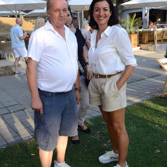 Ein älterer Mann und eine Frau stehen lächelnd und zur Kamera schauend auf einer Rasenfläche. Hinter ihnen sitzen Menschen unter Schirmen an Tischen.