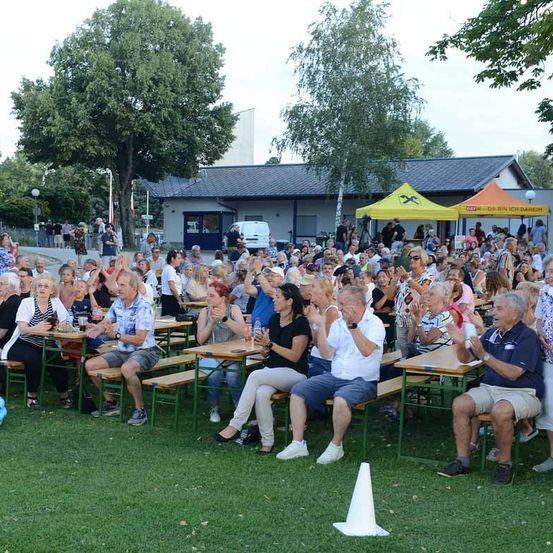 Eine große Gruppe von Menschen sitzt auf Tischen auf einem Rasenfeld, klatscht und lächelt. Die Tische sind in Reihen angeordnet, und im Hintergrund befinden sich Zelte und ein Gebäude.