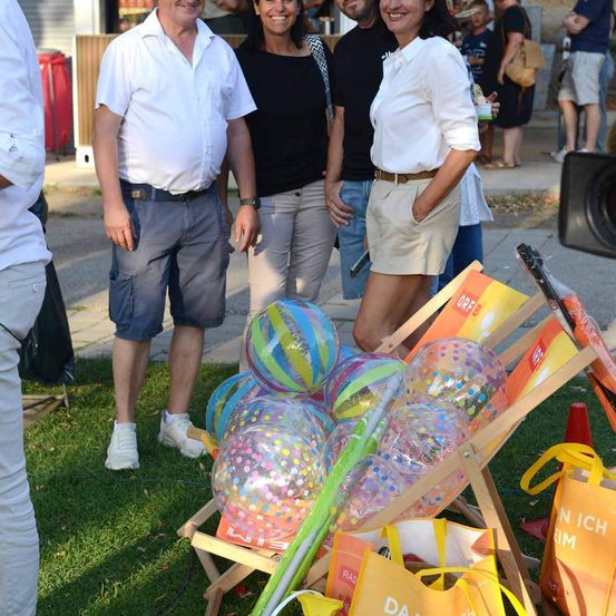 Drei Personen stehen in einem Park mit bunten Luftballons und Taschen vor ihnen. Ein Mann trägt einen Gürtel und Turnschuhe, und eine Frau hat eine Handtasche. Im Hintergrund stehen und gehen Menschen.