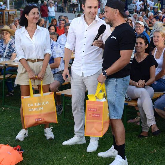 Drei Personen stehen auf einem Rasenfeld, zwei halten orange Taschen mit Text, eine hält ein Mikrofon. Dahinter sitzt eine Menschenmenge auf Bänken.
