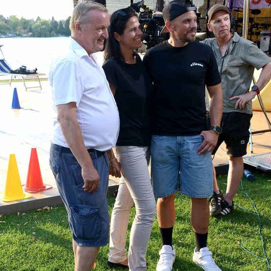 Vier Personen stehen auf einem Rasen und lächeln für ein Foto. Ein Mann in einem weißen Hemd und blauen Shorts steht links. Eine Frau in einem schwarzen Shirt und weißen Hosen steht in der Mitte. Ein Mann in einem schwarzen Shirt und Jeansshorts steht rechts.