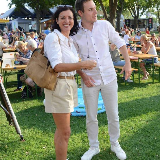 Ein Mann und eine Frau stehen bei einer Outdoor-Veranstaltung, lächeln und posieren für ein Foto. Der Mann trägt ein weißes Hemd und eine Hose, während die Frau ein weißes Oberteil und eine Shorts trägt. Hinter ihnen sitzen Menschen an Tischen, und ein Baum steht im Hintergrund.