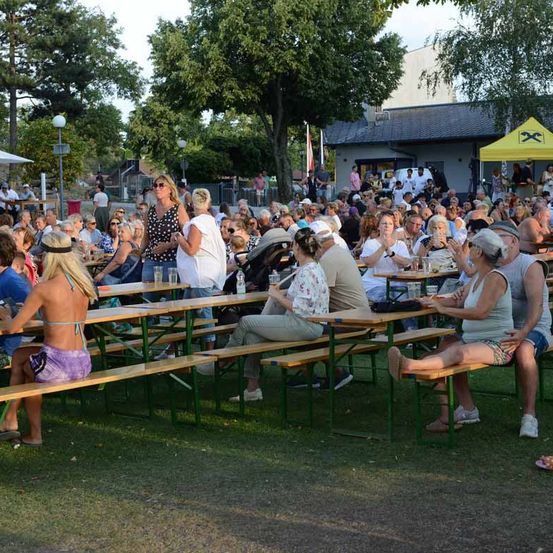 Eine große Menschenmenge sitzt an Picknicktischen im Freien, einige klatschen. Im Hintergrund sind Bäume und ein Gebäude zu sehen.