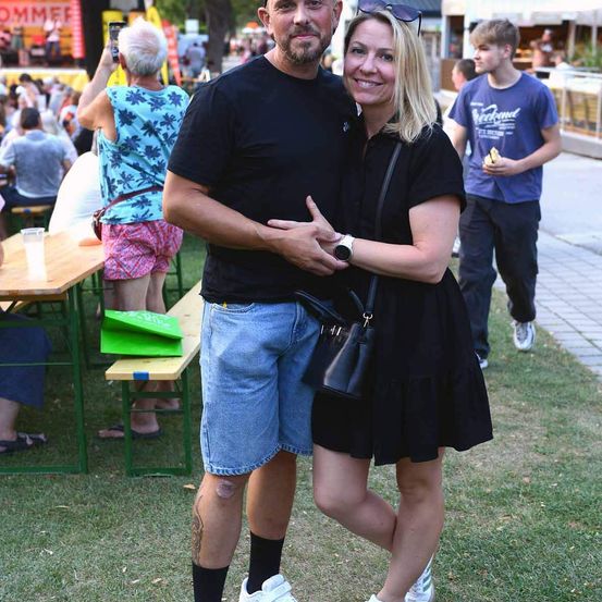 Ein Mann und eine Frau stehen eng beieinander auf einer Veranstaltung im Freien und lächeln für die Kamera. Hinter ihnen sitzen Menschen an Tischen, und eine Bühne ist im Hintergrund zu sehen.