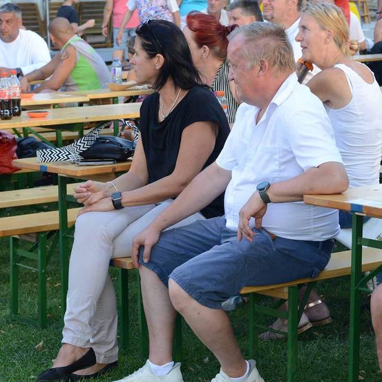 Eine Gruppe von Erwachsenen sitzt auf Bänken bei einer Veranstaltung im Freien, einige unterhalten sich, während andere umherschauen. Flaschen und Teller stehen auf den Tischen.