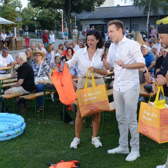 Zwei Personen stehen in einem Außenbereich und halten Taschen in der Hand, während sie in die Kamera sprechen. Ein Publikum sitzt auf Bänken hinter ihnen. Die Umgebung scheint ein öffentliches Ereignis zu sein.