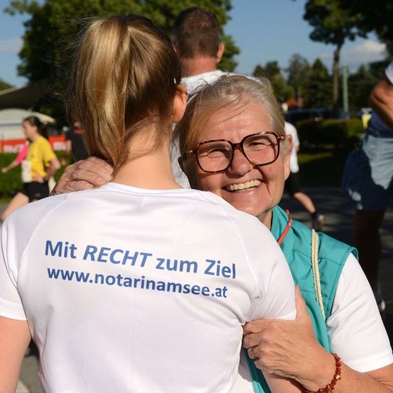 Eine ältere Frau mit Brille lächelt, während sie eine Frau umarmt, die ein weißes Shirt mit blauem Text trägt. Die Frau vor ihr hat einen Pferdeschwanz. Sie stehen auf einem Weg mit anderen Menschen im Hintergrund.