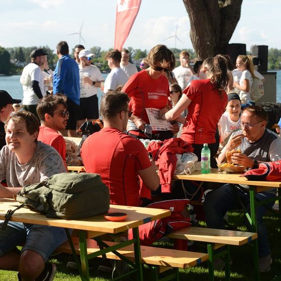 Eine Gruppe von Menschen in roten T-Shirts sitzt an Picknicktischen in einem Park, mit einem See und Windrädern im Hintergrund.