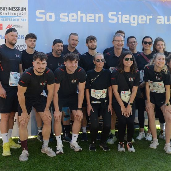 Eine Gruppe von Menschen in passender schwarzer Sportbekleidung mit roten Akzenten posiert für ein Foto vor einem Banner. Jede Person hat eine Nummer auf ihrem Shirt.