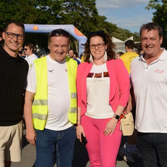 Vier Personen, zwei Männer und zwei Frauen, stehen zusammen im Freien. Sie lächeln und scheinen für ein Foto zu posieren. Der Mann auf der linken Seite trägt eine Brille und ein schwarzes Shirt. Der Mann in der Mitte trägt eine Warnweste und ein weißes Shirt. Die Frau in der Mitte trägt einen rosa Blazer und hält eine Handtasche. Der Mann auf der rechten Seite trägt ein weißes Poloshirt.