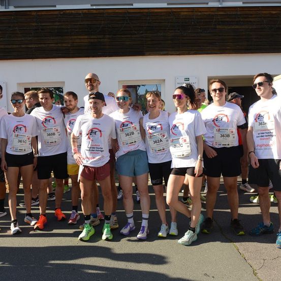 Eine Gruppe von Läufern in passenden weißen T-Shirts mit Nummern und Logos, die vor einem Gebäude stehen und für ein Foto lächeln.