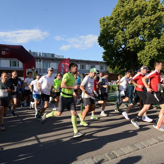 Marathonläufer auf einer Stadtstraße, in verschiedenen farbigen Shirts und Shorts, bei blauem Himmel.