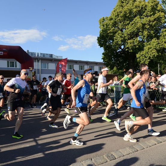 Eine Gruppe von Läufern in Sportbekleidung nimmt an einem Marathon bei sonnigem Wetter teil. Zuschauer säumen die Straße und feuern sie an.