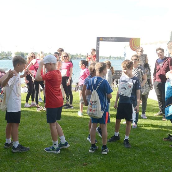 Eine Gruppe von Kindern und Erwachsenen versammelt sich auf einem Rasenfeld, einige tragen Startnummern und Turnschuhe, wahrscheinlich in Vorbereitung auf ein Laufevent. Im Hintergrund ist eine Fotokabine mit einem Burgenland-Schild zu sehen.