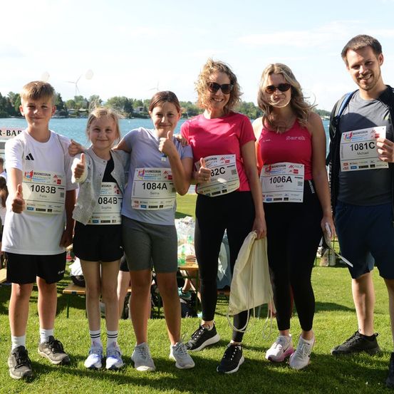 Eine Gruppe von Menschen mit Laufnummern, die auf einem Rasenfeld mit einem See im Hintergrund für ein Foto lächeln.