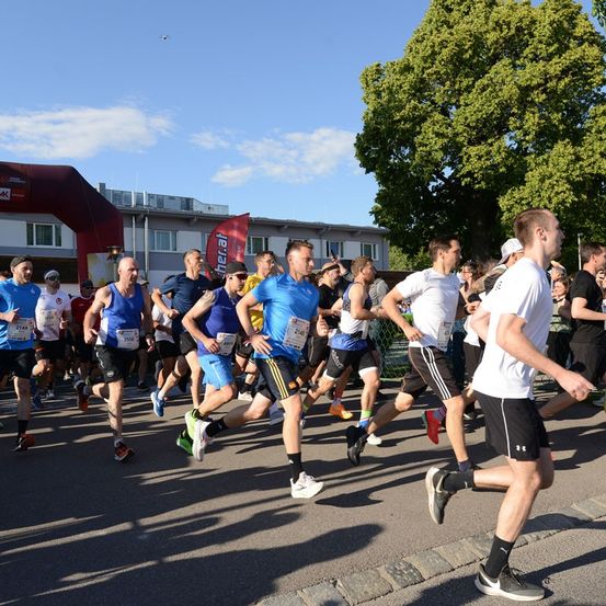 Eine Gruppe von Läufern nimmt an einem Marathon teil und läuft auf einer asphaltierten Straße mit einem Gebäude und einem Baum im Hintergrund.