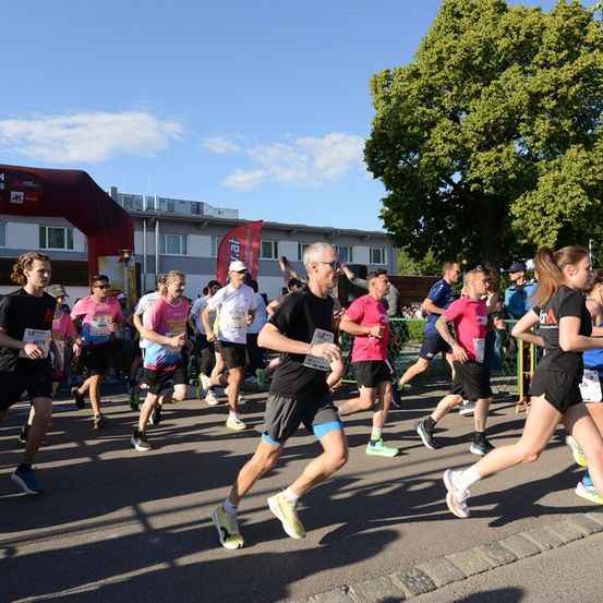Eine Gruppe von Läufern nimmt an einem Marathon teil, läuft auf einer asphaltierten Straße unter einem klaren Himmel, mit einem Baum und einem Gebäude im Hintergrund.